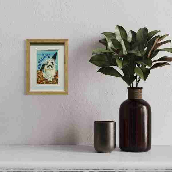 Blue watercolour painting of a rabbit, framed on a neutral wall with houseplants