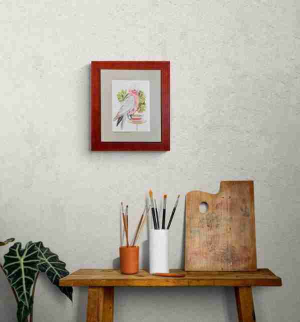 Framed pink galah art on a wall above an artist's side table