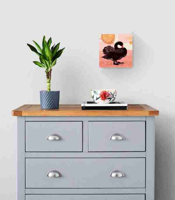 A black swan art print on the wall above a grey cabinet with a plant and floral cup